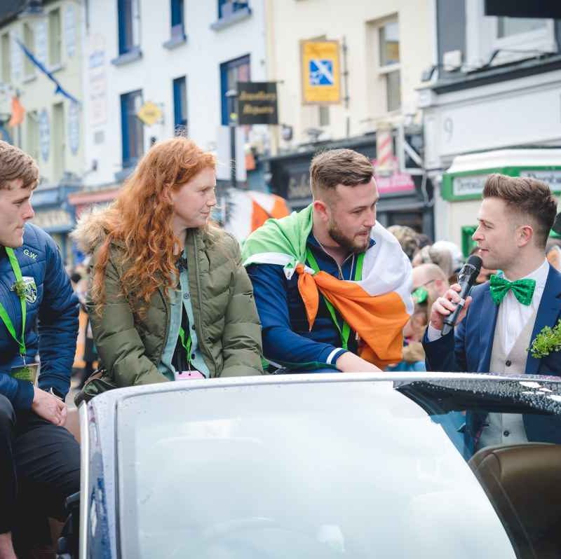 man holding mic asking three people sitting on car a question at St. Patrick's Day Festival Killarney