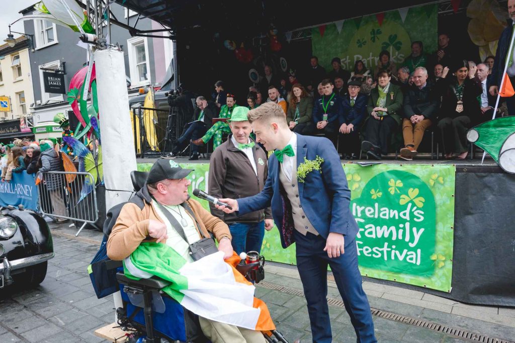man hosting parade holding microphone to men in wheelchair on road in the parade