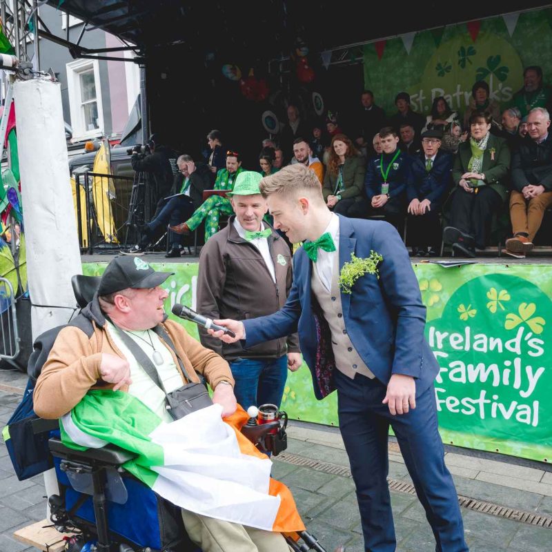 man hosting parade holding microphone to men in wheelchair on road in the parade